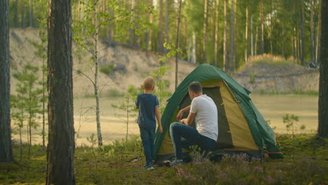 Boy-watches-as-his-father-sets-up-and-assembles-a-tent-in-the-woods-on-the-shore-of-the-lake